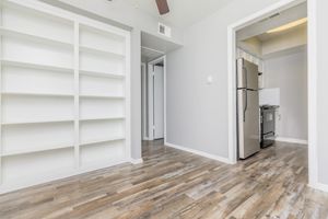An interior view of a modern apartment featuring light gray walls, a white built-in shelf on the left, and a doorway leading to a kitchen area with stainless steel appliances. The flooring is a warm wood tone, and there is a ceiling fan visible in the upper part of the image.