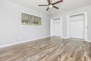 A spacious, empty bedroom featuring light gray walls, a ceiling fan, and wooden flooring. There is a window on one side with blinds, and two open closets with white folding doors on the opposite wall. The room is brightly lit and has a clean, minimalist appearance.