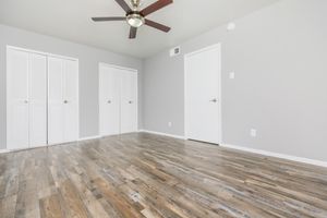 A spacious, well-lit room with light gray walls and wooden flooring. The room features a ceiling fan and two sets of double-door closets. There is a single door on one wall, and the overall design conveys a modern, minimalist aesthetic.