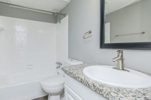 A clean, modern bathroom featuring a white bathtub and shower combination, a toilet, and a granite countertop with a sink. The walls are painted gray, and there is a large mirror above the sink. A towel bar and a wall-mounted fixture are visible. The overall design is simple and stylish.