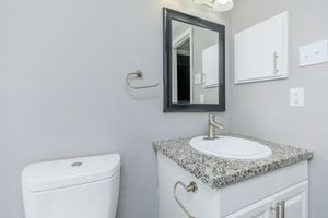 A modern bathroom featuring a gray wall, a white vanity with a granite countertop and a circular sink, a wall-mounted mirror with a black frame, a light fixture above, a toilet, and a towel bar. There is also a small cabinet on the wall for storage.