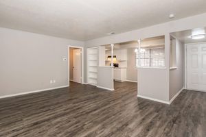 A spacious, modern living area with light-colored walls and dark wood-style flooring. The room features a doorway leading to another room, an open kitchen area with white cabinetry, and large windows with blinds. The overall ambiance is bright and inviting, ideal for comfortable living.