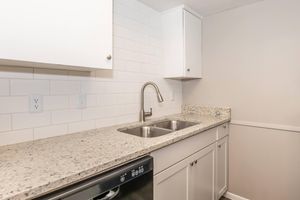 Modern kitchen featuring a double sink with a sleek faucet, granite countertops, and white cabinetry. The backsplash consists of white tiles, and there is a built-in dishwasher beneath the counter. The walls are painted in a soft gray, creating a clean and contemporary look.