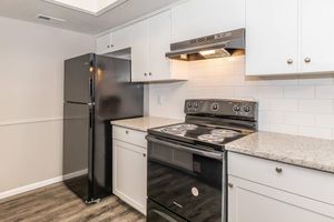 A modern kitchen featuring a black refrigerator, a black oven with an electric cooktop, and a sleek vent hood. The cabinets are white, and the countertops are speckled granite. The flooring is dark wood laminate, creating a contemporary and inviting atmosphere.