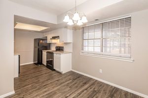 A modern kitchen with a refrigerator, stove, and white cabinetry. The walls are a neutral color, and there's a window with blinds letting in natural light. The flooring is a wood-like laminate, and a light fixture hangs from the ceiling. The space appears clean and well-organized.