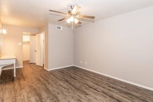 A spacious, empty living room with light gray walls and a ceiling fan. The floor has wooden laminate, and there is a white table against one wall. A doorway leads to another room, while the opposite side has a door opening, suggesting additional areas. Natural light is present, enhancing the room's brightness.