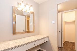 A well-lit vanity area featuring a large mirror with three light fixtures above, a countertop made of granite or a similar material, and a white cabinet below. To the right, there's an open door leading to a small closet with a hanging rod, set against a neutral wall color and dark laminate flooring.