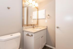 A clean and modern bathroom featuring a white toilet, a light-colored granite countertop with a sink, a large mirror above the sink, and stylish lighting fixtures. The walls are painted a soft gray, and there are towel bars and a small cabinet for storage.