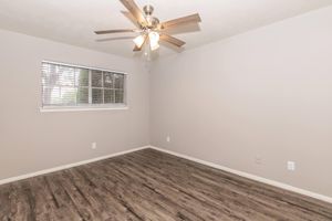 A light and airy empty room featuring a ceiling fan with wooden blades, a window with blinds allowing natural light, and light gray walls. The floor is covered with laminate wood-style flooring, creating a warm and inviting atmosphere.