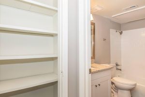 A modern bathroom featuring a white toilet, granite countertop, and a shower/tub combination. Adjacent to the bathroom is a shelf unit with empty white shelves, showcasing a clean and minimalistic design. The walls are painted in a soft gray color, enhancing the overall brightness of the space.