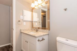 A modern bathroom featuring a white vanity with a granite countertop, a mirror above the sink, and a wall-mounted towel holder. A white toilet is visible on the right side, with neutral-colored walls and a light fixture above the mirror. The flooring is dark wood.