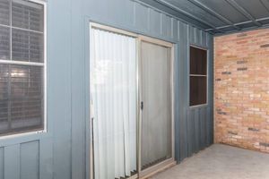 A view of a patio area featuring a sliding glass door with vertical blinds, alongside two windows—one with a grid pattern—and a wall with a brick finish. The exterior is painted in a muted blue color, creating a contrasting backdrop against the brick. The scene is well-lit, suggesting a daytime setting.