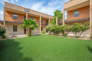A well-maintained courtyard featuring green synthetic grass, palm trees, and flowering bushes. Two levels of apartment buildings surround the space, with several balconies visible. The sky is bright with scattered clouds, creating a pleasant, inviting atmosphere.