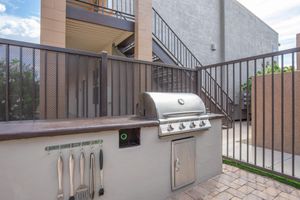 A modern outdoor BBQ grill is mounted on a granite countertop, accompanied by grilling utensils hanging on a wall. The area is enclosed by a metal fence, with a staircase and building in the background. Lush greenery is visible along the fence, creating a welcoming outdoor cooking space.