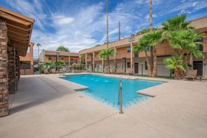 A sunny pool area surrounded by palm trees and lounge chairs, with a two-story building in the background. The pool reflects the blue sky and is located in a well-maintained courtyard, creating a relaxing atmosphere.