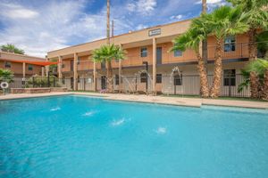 A swimming pool surrounded by palm trees and lounge chairs, with a multi-story building in the background. The building features balconies and is painted in warm tones. Water splashes can be seen in the pool, and a clear blue sky is overhead.