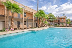 A sunny outdoor pool area surrounded by palm trees and lounge chairs, with a two-story building in the background. The building features balconies, and there are decorative plants and a water fountain near the pool. Bright blue skies with some clouds complete the scene.