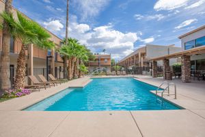 A sunny outdoor pool area surrounded by palm trees, lounge chairs, and apartment buildings. The clear blue water of the pool reflects a bright sky with scattered clouds. A landscaped area with flowers enhances the inviting atmosphere for relaxation and leisure.