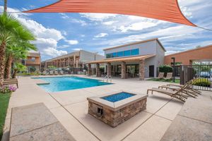 A well-maintained outdoor pool area featuring a clear blue pool, sun loungers, and a covered patio. A modern building is in the background, complemented by palm trees and shaded by a large fabric canopy. The sky is partly cloudy, creating a bright, inviting atmosphere.