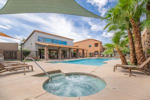 A well-lit outdoor pool area features a clear blue swimming pool surrounded by lounge chairs. There is a spa/jacuzzi on one side, shaded by large sail shades. Palm trees and modern buildings create a relaxed atmosphere, with a clear sky and fluffy clouds in the background.