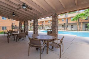 A shaded patio area with multiple tables and chairs overlooking a swimming pool. The pool area is surrounded by palm trees and features a few lounge chairs. The architecture includes columns made of stone and a view of several apartment balconies in the background.