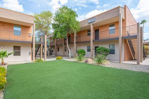 A view of a multi-story residential building with a modern design, featuring several balconies and staircases. The foreground includes a well-maintained green lawn with shrubs and landscaping, while the background shows a partly cloudy sky. The setting is inviting and neatly organized.