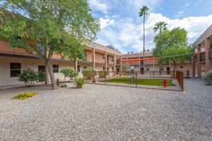 A courtyard surrounded by two-story buildings, featuring gravel ground, small bushes, and a few trees. In the center, there is a patch of green grass enclosed by a fence. A fire hydrant can be seen nearby, and the sky is partly cloudy with trees and palm trees in the background.