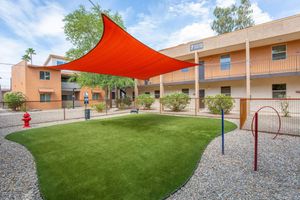 A spacious outdoor area featuring artificial grass, shaded by a large orange canopy. Surrounding the greenery are small shrubs and a fire hydrant. In the background, there are two-story buildings with balconies and an accessible pathway. Ideal for relaxation and social gatherings.