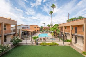 A view of a residential area featuring two-story buildings arranged around a central pool. The pool is enclosed with a black fence, surrounded by palm trees and landscaped greenery. The sky is partly cloudy, adding to the serene atmosphere of the community.