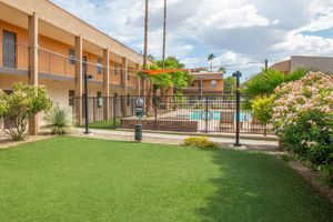 View of a hotel's outdoor area featuring a green lawn, tropical plants, and a fenced swimming pool. The building includes two levels of guest rooms with balconies. A volleyball net is set up on the lawn, and palm trees can be seen in the background under a partly sunny sky.