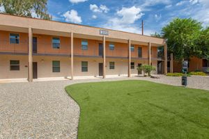 A well-maintained courtyard of a multi-story apartment building featuring a green lawn, gravel pathways, and a sign indicating unit number 3. The building has multiple balconies and large windows, with trees and shrubbery enhancing the outdoor space under a partly cloudy blue sky.