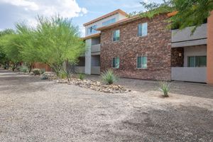 Exterior view of a residential building featuring a mix of stone and stucco façades. The building has two levels, with several windows and a flat roof. Surrounding the structure are desert plants and gravel landscaping, with a clear blue sky above and green foliage nearby.