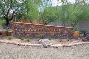 A stone monument sign for "Casa Presidio Apartment Homes," featuring the year "2002" and surrounded by plants and decorative rocks. The background includes green trees and a portion of a building.
