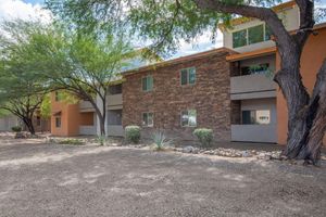 A peaceful exterior view of an apartment community featuring earthy-toned brick and stucco buildings surrounded by trees and desert landscaping. The area has a gravel path and rocks, creating a natural setting. The sky is partly cloudy with a hint of sunlight illuminating the scene.