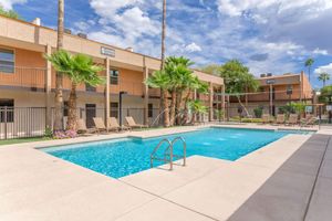 A view of a swimming pool surrounded by palm trees and lounge chairs, with a two-story building in the background. The sky is partly cloudy, indicating a pleasant day. The pool area is enclosed with a fence, creating a private and inviting atmosphere.