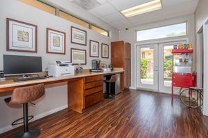 A bright modern office space featuring a wooden desk with a computer and printer, a coffee maker, and decorative framed prints on the walls. There's a small red cart with colorful snacks, and a door leading outside, providing a view of a patio area. The floor is covered in dark wood laminate.