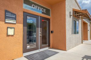 Entrance of a leasing office with a sign reading "OFFICE" and "OPEN." The exterior features a textured orange wall, a mailbox, and a notice board beside the door. Clear blue skies are visible above, adding to the welcoming atmosphere.