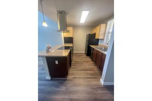 Modern kitchen featuring wooden cabinetry, stainless steel appliances, and a spacious countertop. The layout includes an island with bar seating, and large windows allowing natural light. The flooring is wood-like, enhancing the contemporary feel of the space.