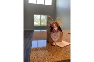 A decorative gift box with a heart-shaped cutout and a bow sits on a granite countertop in a spacious room with large windows. A handwritten card is placed next to the box, adding a personal touch. The overall setting suggests a warm, welcoming atmosphere.