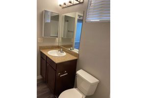 A small bathroom featuring a beige countertop sink with a mirror above and a wooden cabinet beneath. To the right, there is a white toilet and a light fixture above the sink. The walls are painted in a light color, and there are partially closed blinds on a window allowing natural light in.
