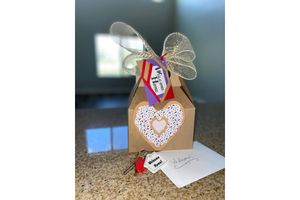A beautifully wrapped gift box made of kraft paper, adorned with a decorative lace heart cutout and a large bow made of natural mesh. It features colorful tags, one reading "Welcome" and a card with a handwritten note. A set of keys rests beside the box on a granite countertop, with soft natural light in the background.