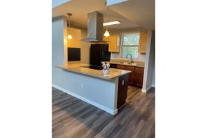 A modern kitchen featuring a central island with a granite countertop, dark wooden cabinets, stainless steel appliances, and a black fridge. Pendant lights hang above the island, and a window provides natural light. The floor is finished with light-colored wood laminate.