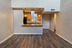 Open-concept living space featuring a modern kitchen with a central island and pendant lights. Light wood cabinetry contrasts with dark appliances. The area has wood-like flooring and neutral-colored walls, with natural light coming from a window. A doorway leading to another room is visible in the background.
