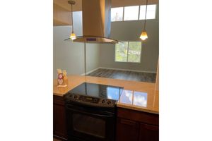 A modern kitchen featuring a sleek black stove and a hood above. The countertop is light-colored and there are two pendant lights hanging down. In the background, a spacious living area with large windows allows natural light to filter in, showcasing freshly painted walls and flooring.