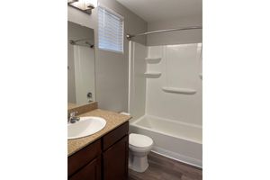 A clean, small bathroom featuring a bathtub with a shower curtain, a white toilet, and a sink with a granite countertop. The mirror above the sink reflects the room, and there is a window with blinds allowing natural light. The wall color is light, adding to the spacious feel.
