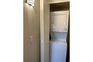 A stacked washer and dryer unit situated in a small laundry nook, with a light fixture on the wall beside it. The unit is white and enclosed by a door that is partially ajar, revealing the appliances inside a narrow space. The wall is painted a light neutral color.