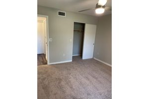 A view of a small, well-lit bedroom featuring beige carpet and light gray walls. There’s a ceiling fan overhead, a closet with sliding doors on the right, and a doorway leading to another room on the left. The space appears tidy and freshly painted, creating a cozy ambiance.