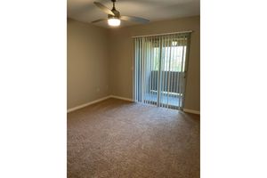 An empty room with light beige carpet and light gray walls. A ceiling fan is mounted on the ceiling, and there is a large sliding glass door with vertical blinds that leads to a balcony or outdoor area. Natural light enters through the door, illuminating the space.