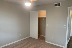 A clean and empty room with light gray walls and a ceiling fan. There is a beige carpeted floor and a white closet with an open sliding door, revealing a simple hanging rod inside. The room has a bright, minimalist feel.
