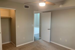Interior view of a room featuring beige walls and carpeted floors. There's a ceiling fan, an open door leading to another room, and a closet space visible on the left. Natural light enters the room from the doorway, highlighting the spaciousness of the area.